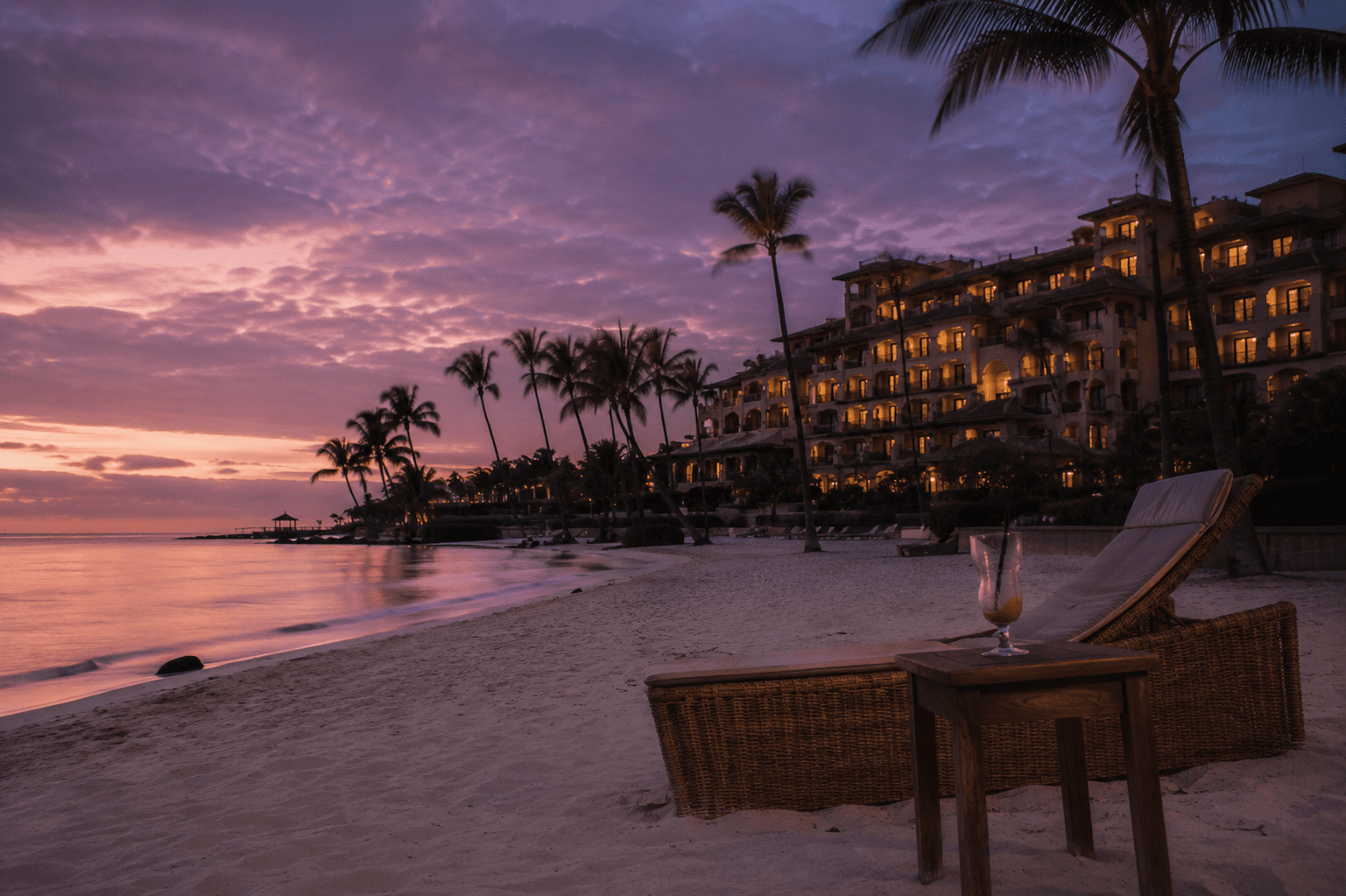 An empty wicker beach lounger and a half-finished cocktail on a Caribbean beach at twilight, a luxury resort glowing in the background