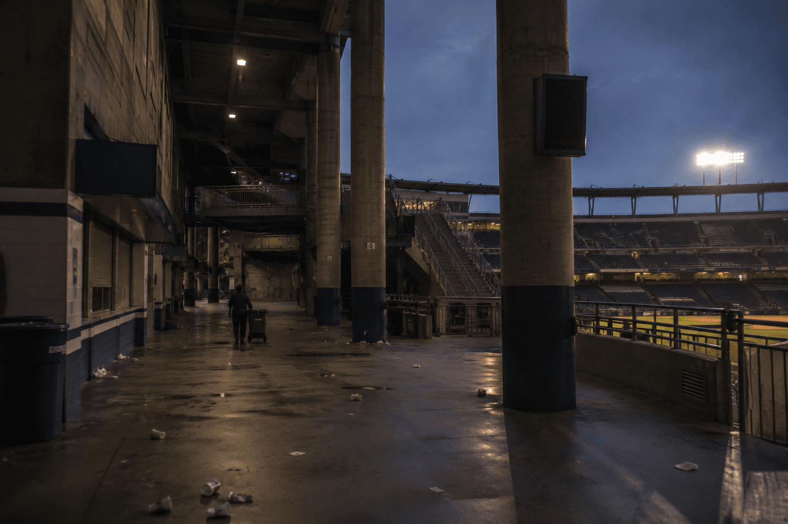The vast empty concourse of an American sports stadium at twilight, scattered debris from the day's crowd, a single janitor in the distance